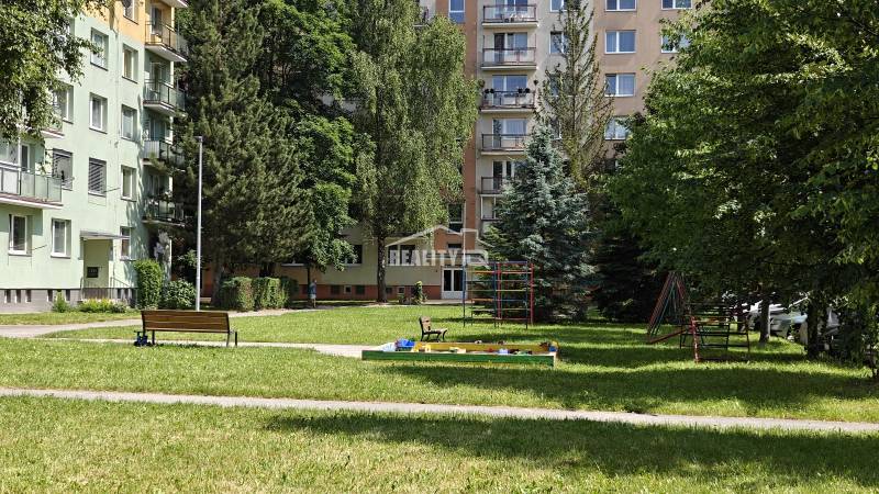 Detské ihrisko medzi bytovkami v Žiline, obklopené stromami a lavičkami.